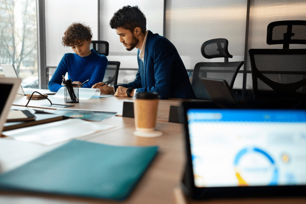 Office colleagues collaborate using a tablet and laptop, surrounded by notes and personal items.