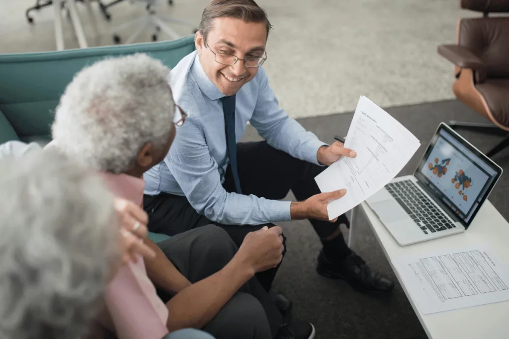 Man in a suit discussing paperwork with elderly clients, with a laptop showing charts on a table.