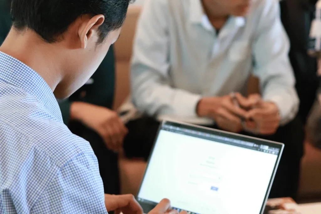 Focused individual using laptop in a collaborative group setting with visible screen.