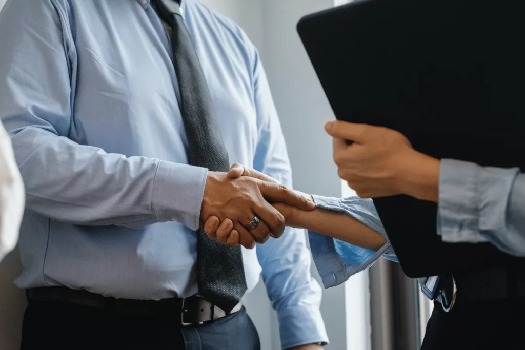 Two people in formal attire greeting each other with a handshake in front of a bright window.