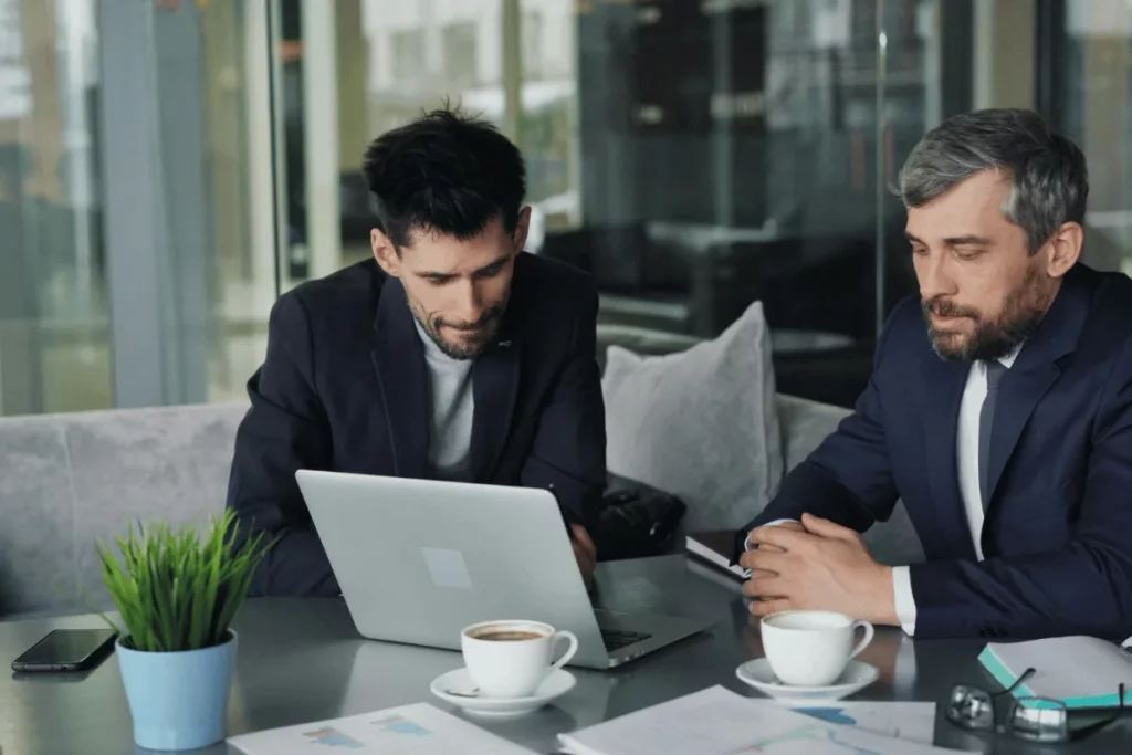 Two businessmen reviewing data on a laptop during a meeting with coffee cups and documents on the table.