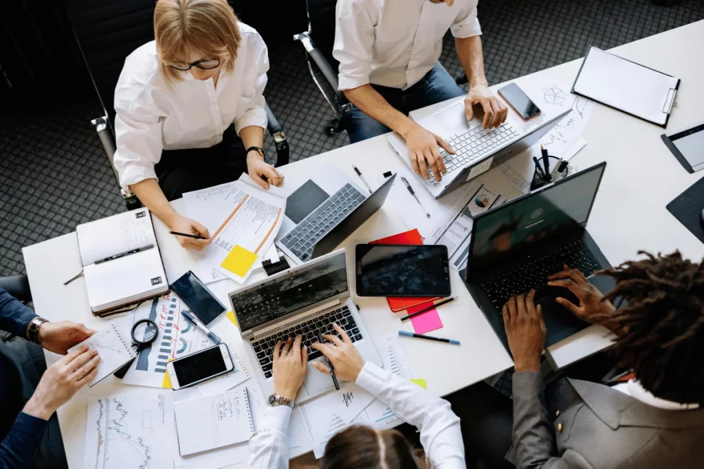 Team of professionals working around a table with laptops, documents, and smartphones.