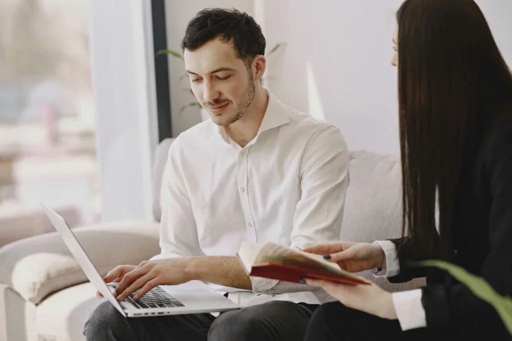 Two colleagues discussing work, with one using a laptop and the other holding a book.