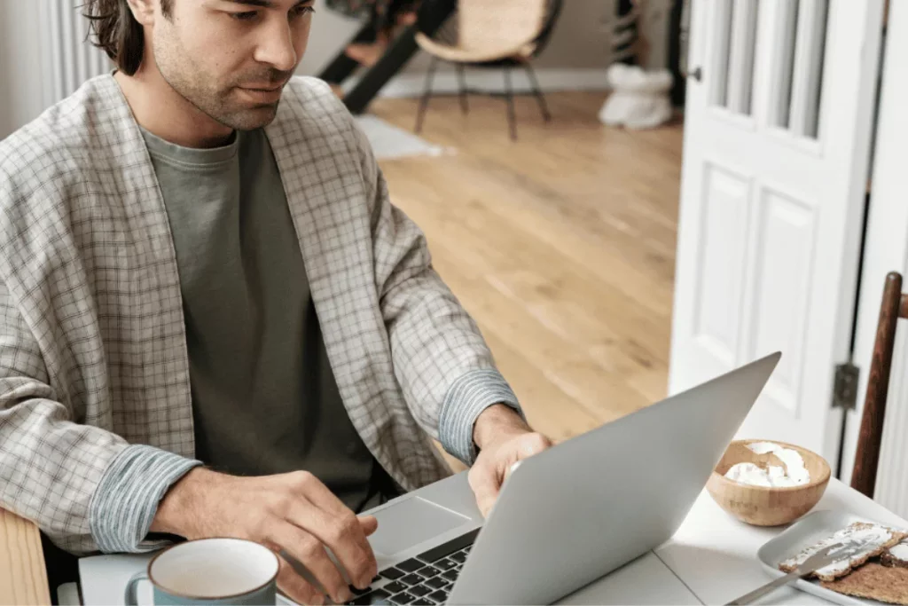 A man in a plaid robe working on a laptop with a coffee cup and a bowl of yogurt on the table in a cozy home setting.