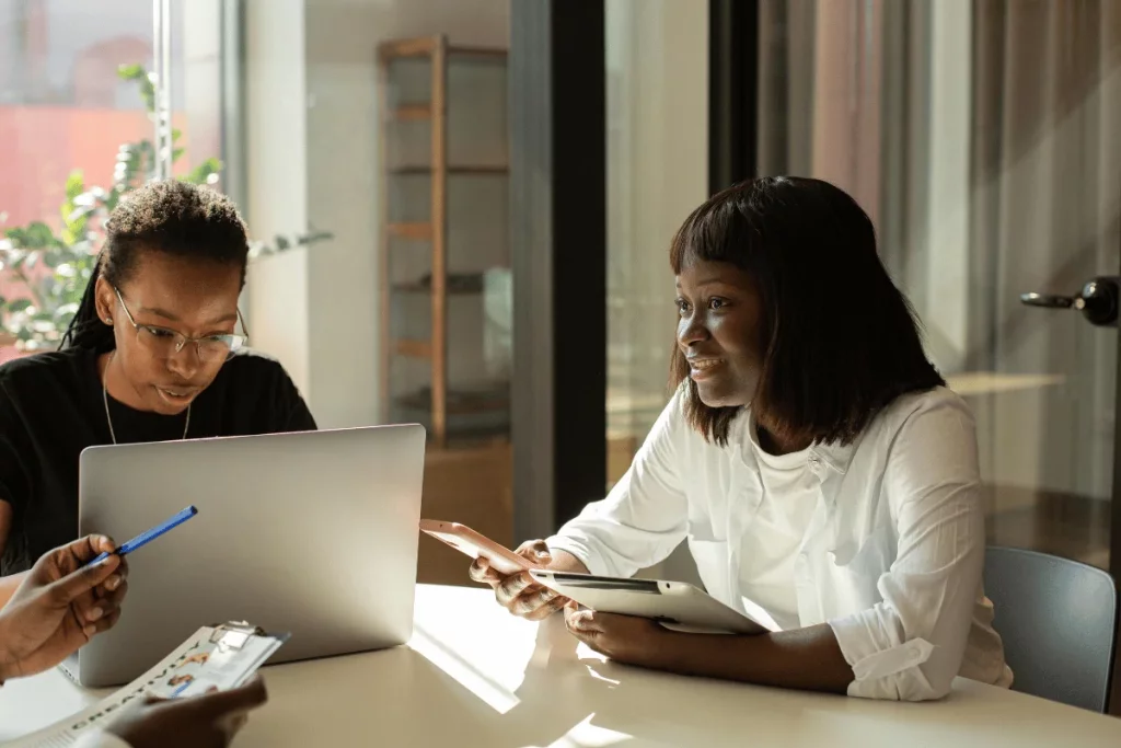 Two women collaborate around a table in a modern office, one on a laptop and the other holding a tablet.