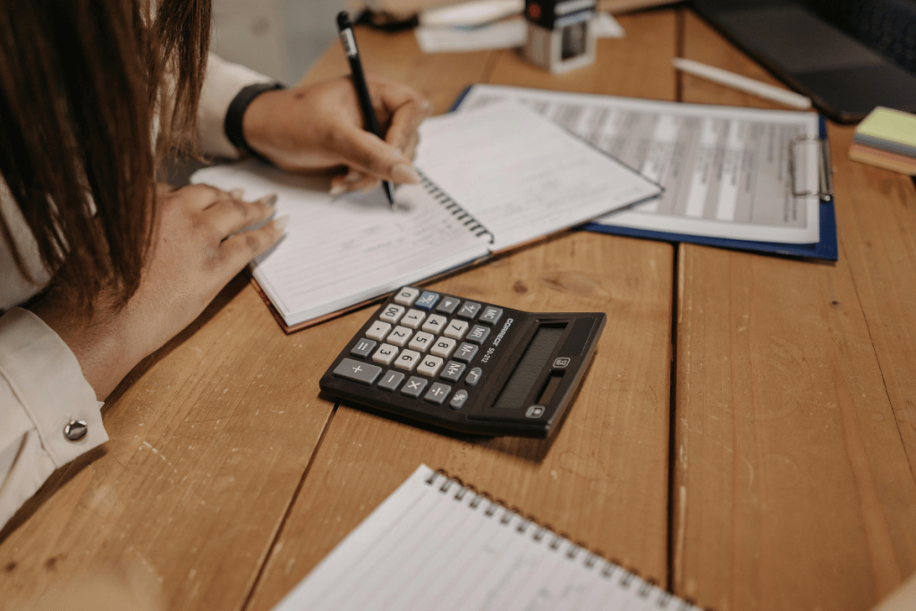 Person jotting down notes with a pen beside a calculator on a wooden table.