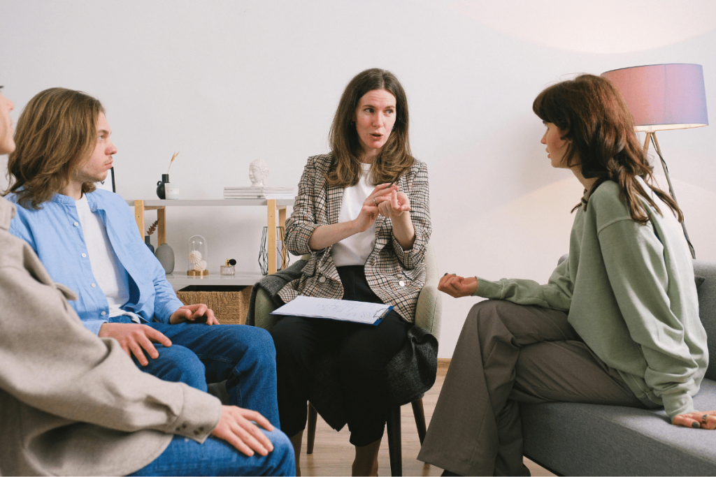 Three individuals engaged in a serious conversation, with one woman gesturing while speaking to the other two seated across from her.
