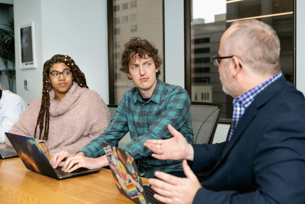 A diverse group of people engage in a discussion around a conference table with laptops in a modern office setting.