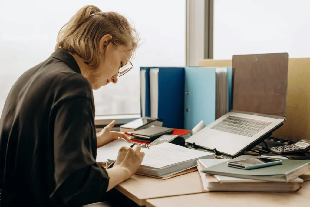 Woman writing in a notebook while working at a desk with a laptop and files in an office setting.