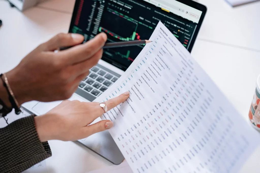 Hands point to a printed financial report while discussing data on a laptop screen displaying stock charts and market information.