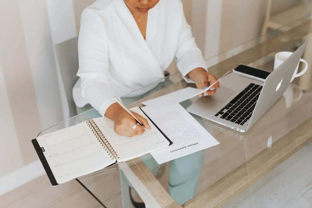Woman taking notes in a planner while reviewing papers and working on a laptop at her desk.