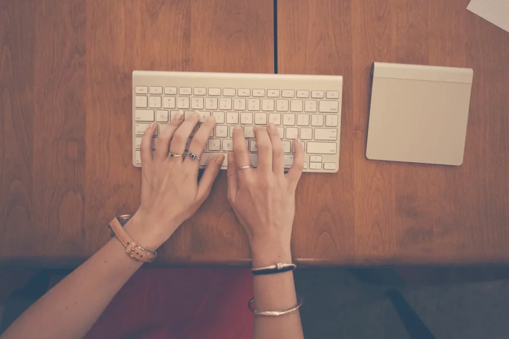 Close-up of female hands with jewelry typing on an Apple keyboard at a desk.