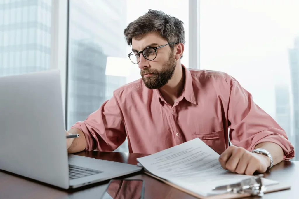 Man in glasses working on a laptop while reviewing documents in a modern office setting.