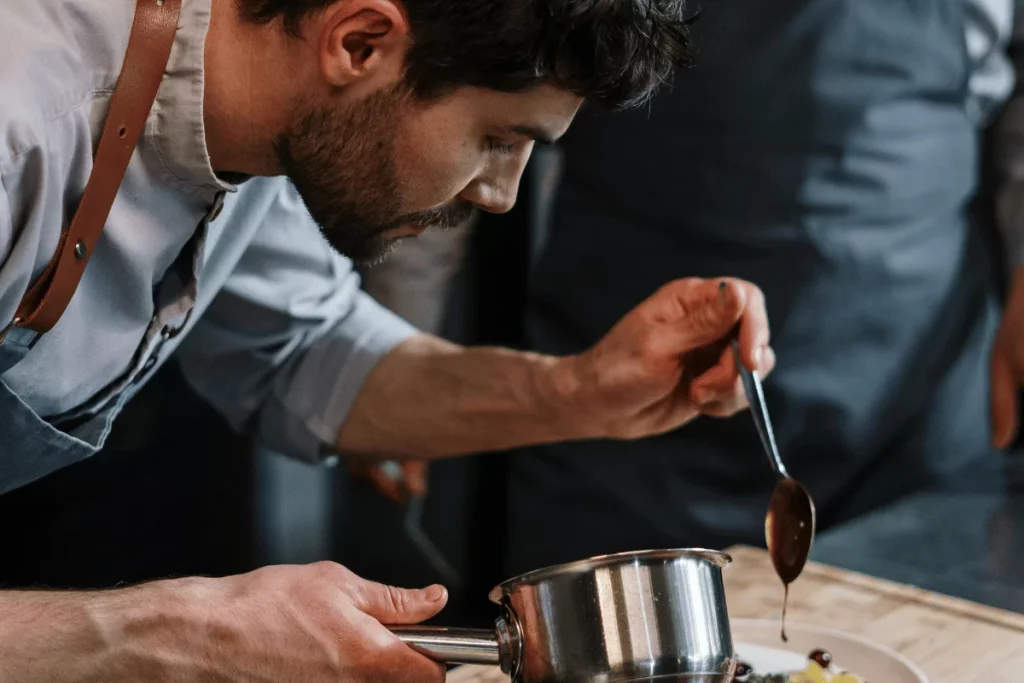Chef in a blue shirt drizzling sauce over a dish from a metal pot.