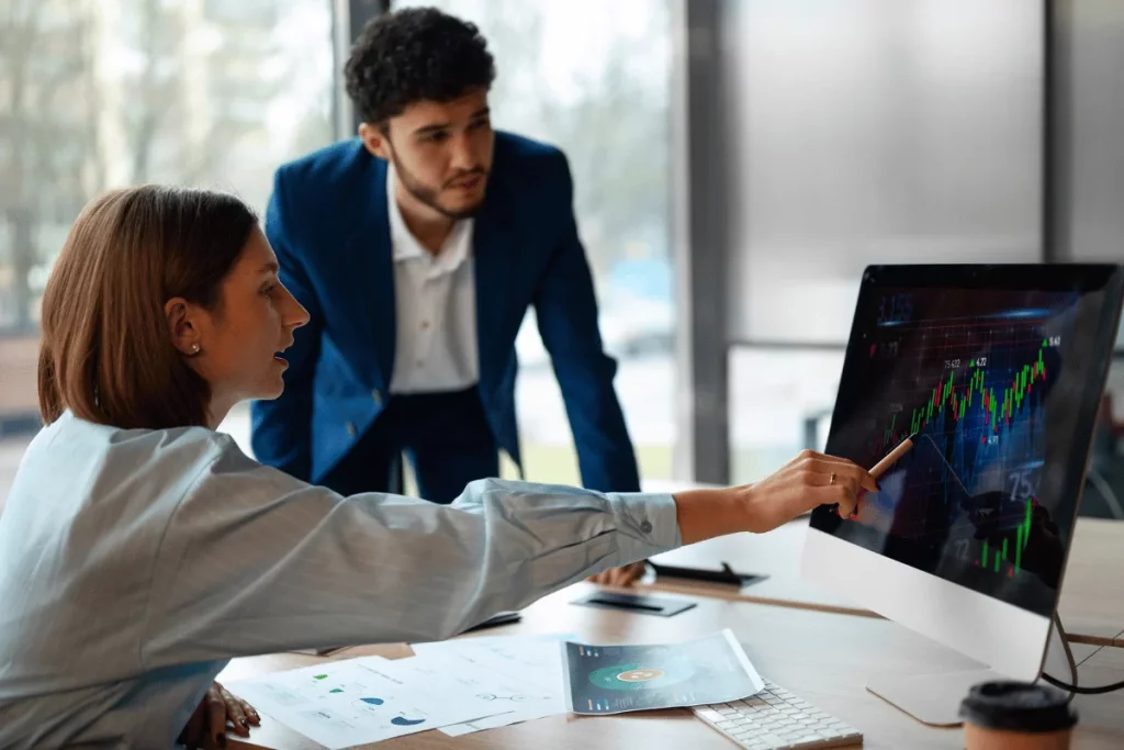 A professional duo analyzes data on a computer screen, with graphs and charts visible on the display and documents scattered on the desk.