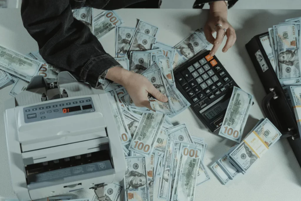 A person counting cash near a calculator and a money counter on a table.