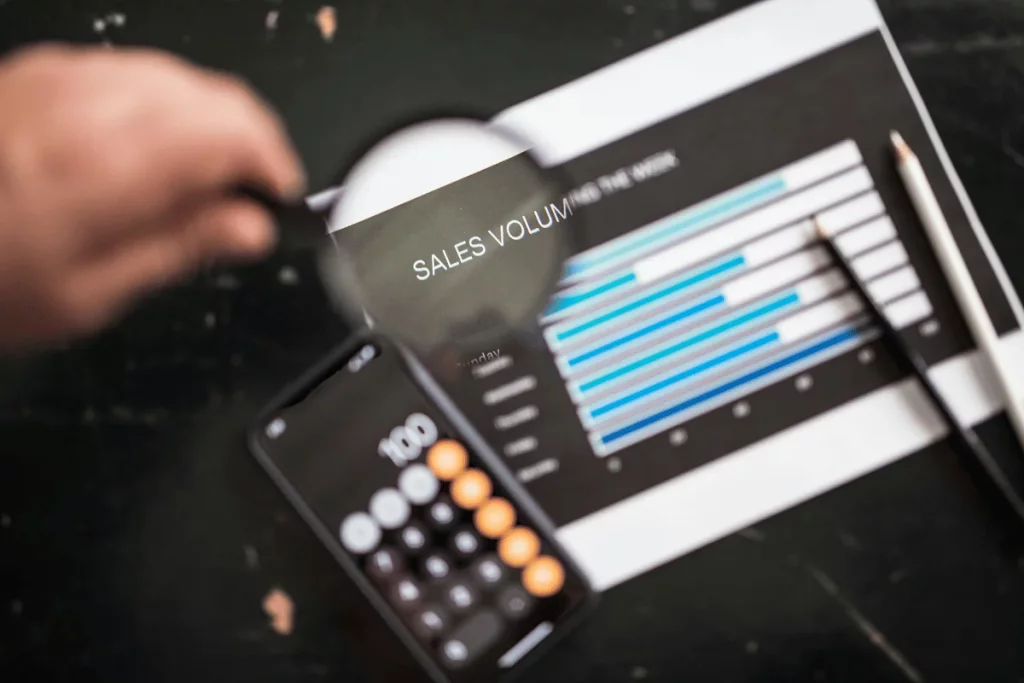 Person using a magnifying glass to review a sales volume chart with a smartphone and pencil on the table.