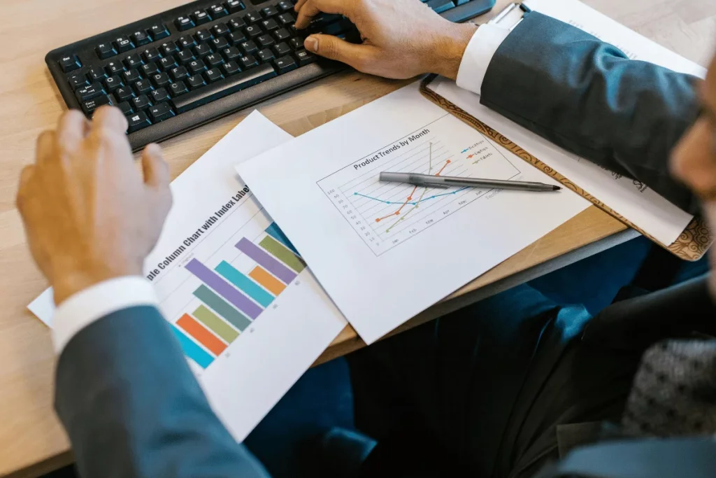 A person in a suit reviewing a column chart and a product trend line chart on paper while using a computer.
