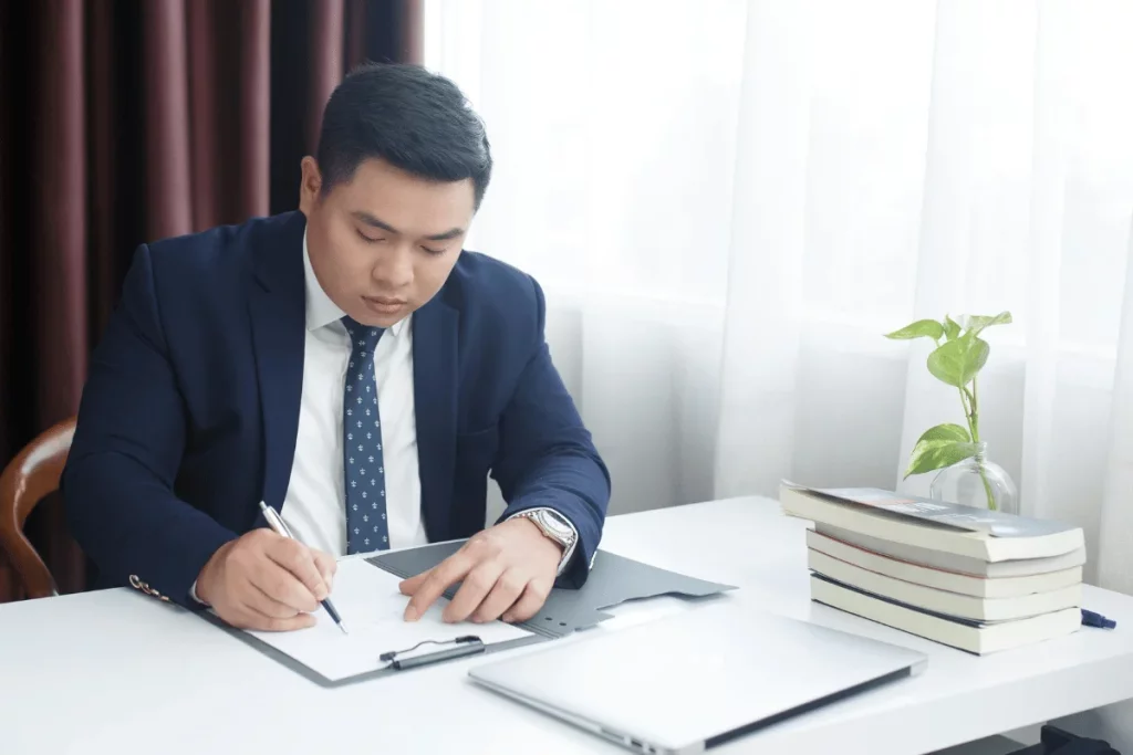 Man in a suit writing on a document at a desk with a laptop, books, and a plant.