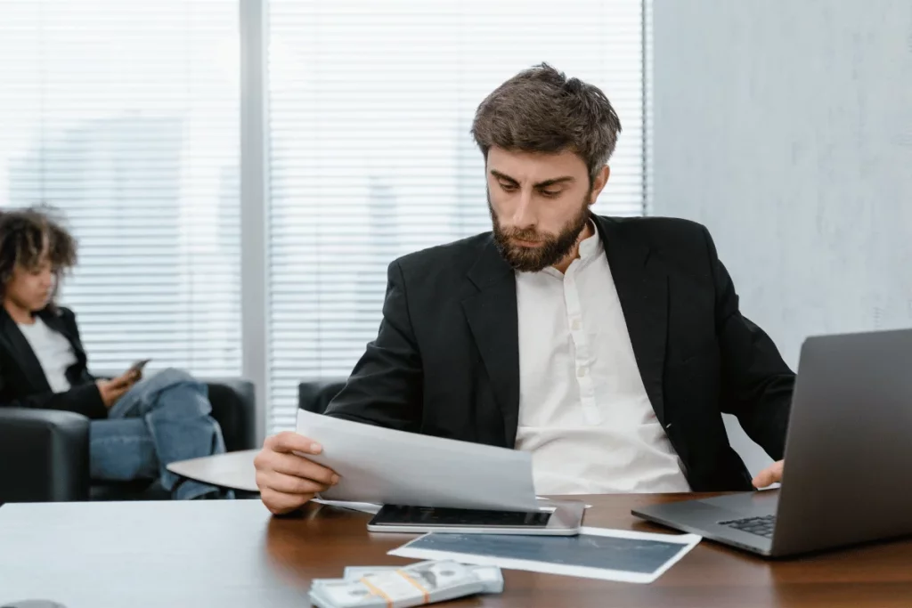 Man in suit reviewing documents with laptop and money on desk.