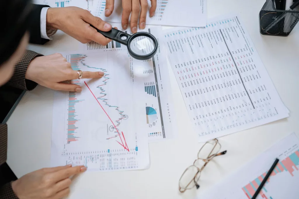 Hands analyzing charts with magnifying glass on a desk with glasses.