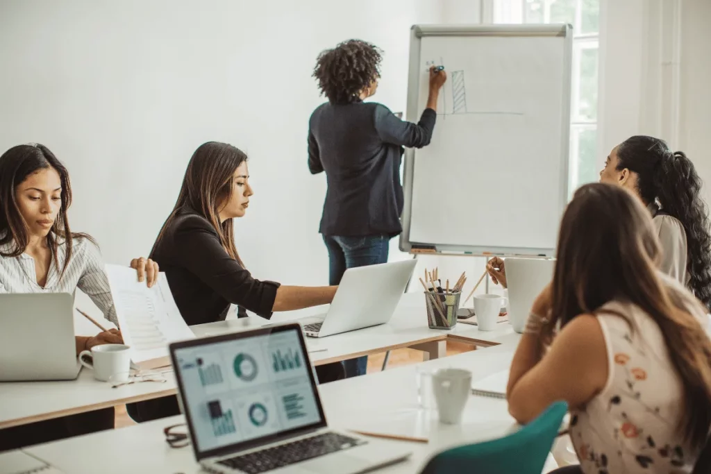 A person presenting on a whiteboard in a meeting room with attendees at a table with laptops.
