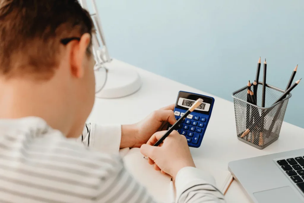 Person calculating with a blue calculator at a desk, surrounded by pencils and a laptop.