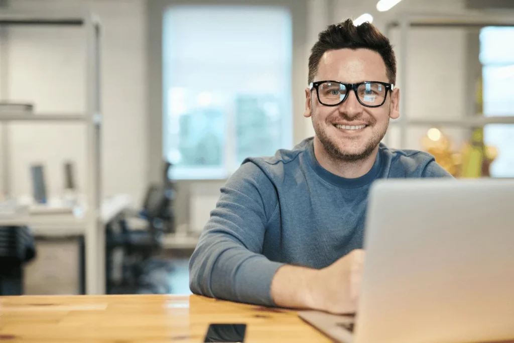 A smiling man in glasses working on his laptop in a modern office.