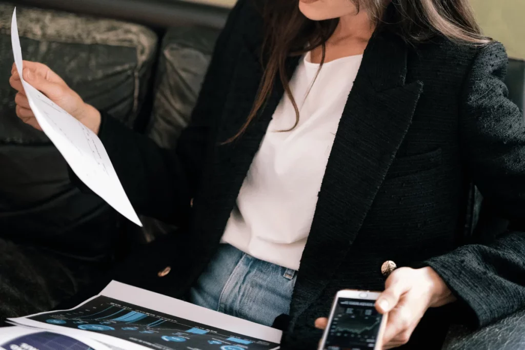 A focused individual in a blazer sits while reading a paper and checking her smartphone.