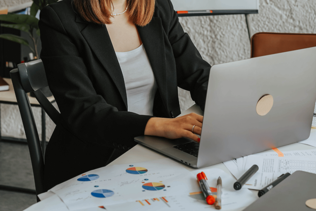 Professional in blazer typing on laptop near financial pie charts.