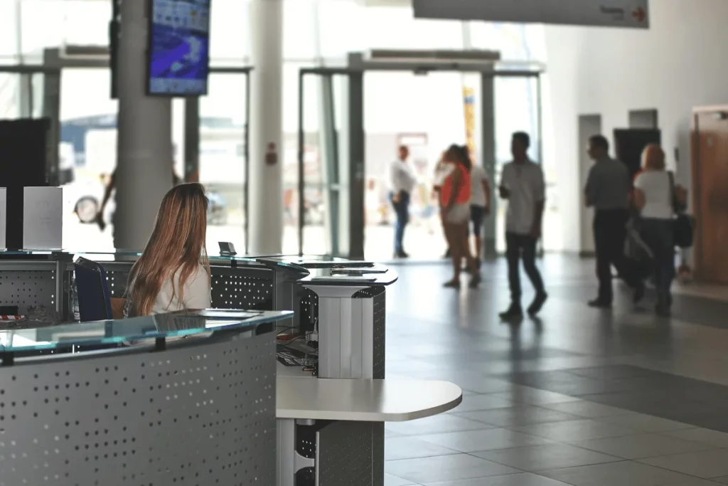 A reception area in a bright, modern building, with a view of people walking towards the entrance and a digital screen displaying information.