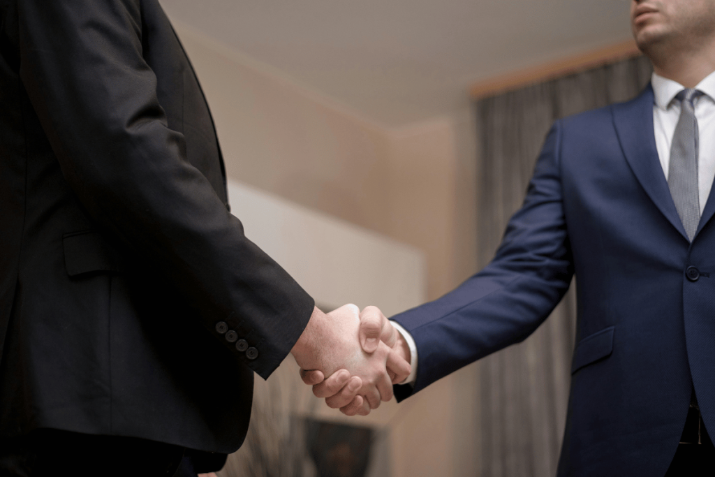 Two professional businessmen in suits engaging in a firm handshake in an office setting.