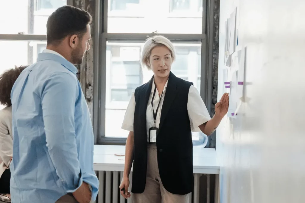 Businesswoman explaining a strategy to a colleague while pointing at charts on a whiteboard.