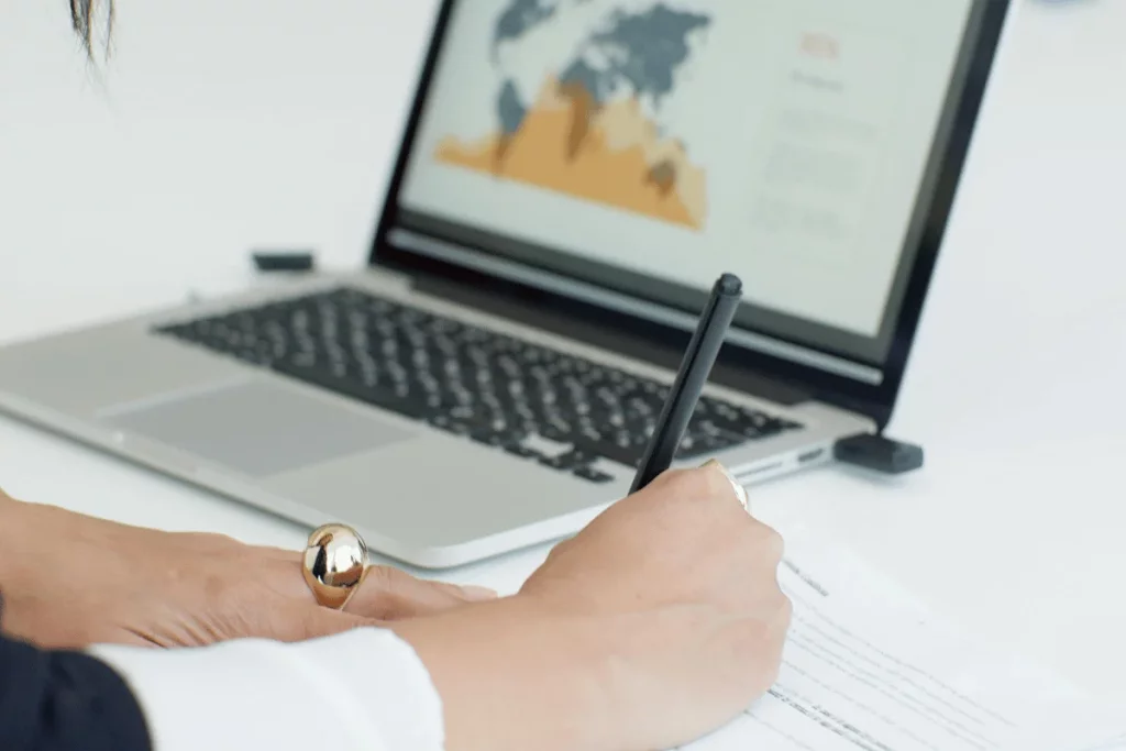 Close-up of a person writing while working on a laptop displaying a data chart.