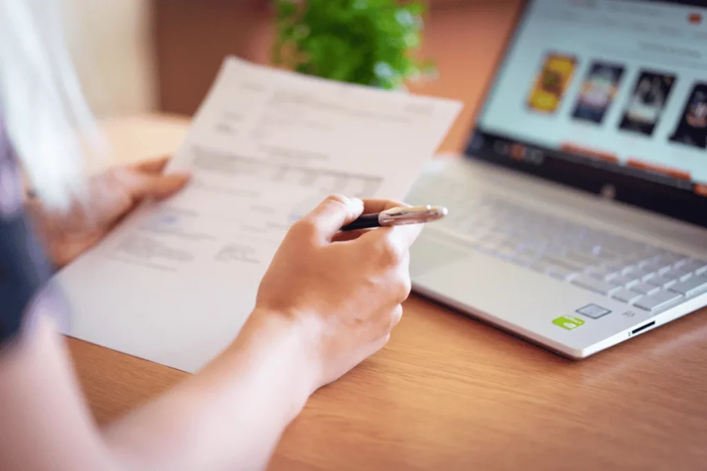 Hands holding a pen, examining a printed document, with a laptop displaying an online store on the desk.