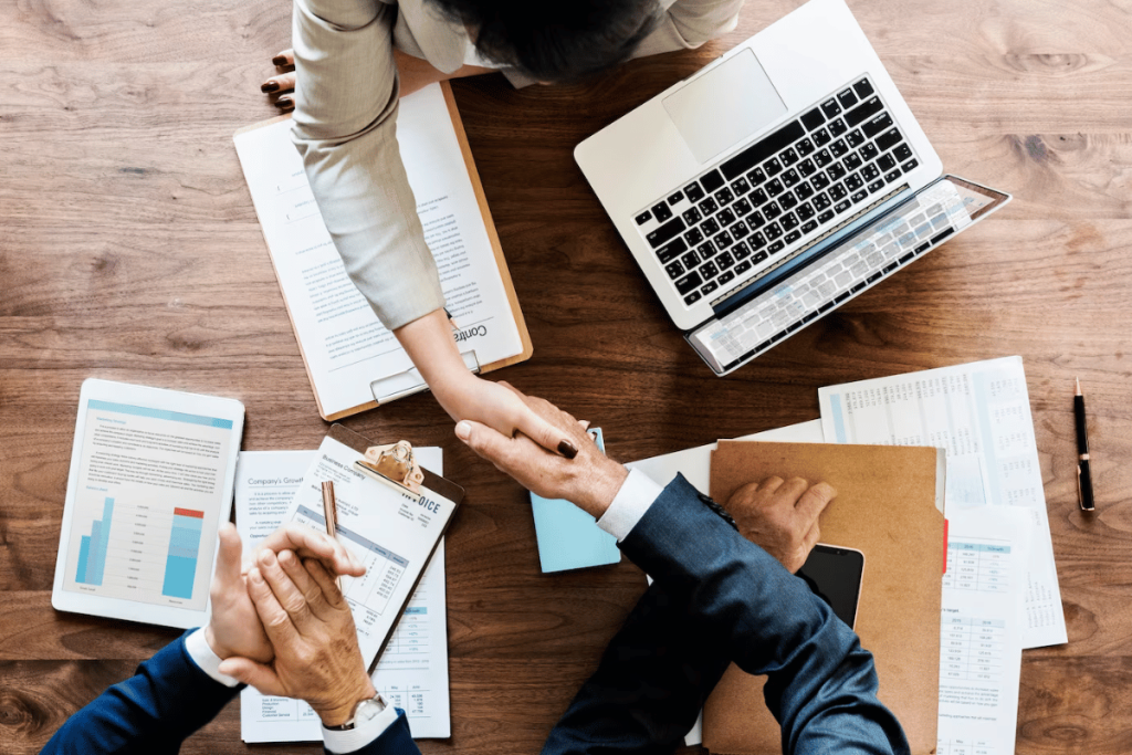 Business people shaking hands at a table with papers and laptops.