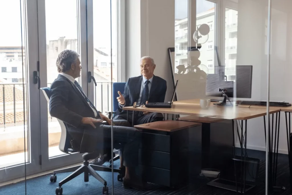 Two businessmen in suits having a conversation in a modern office with large windows.