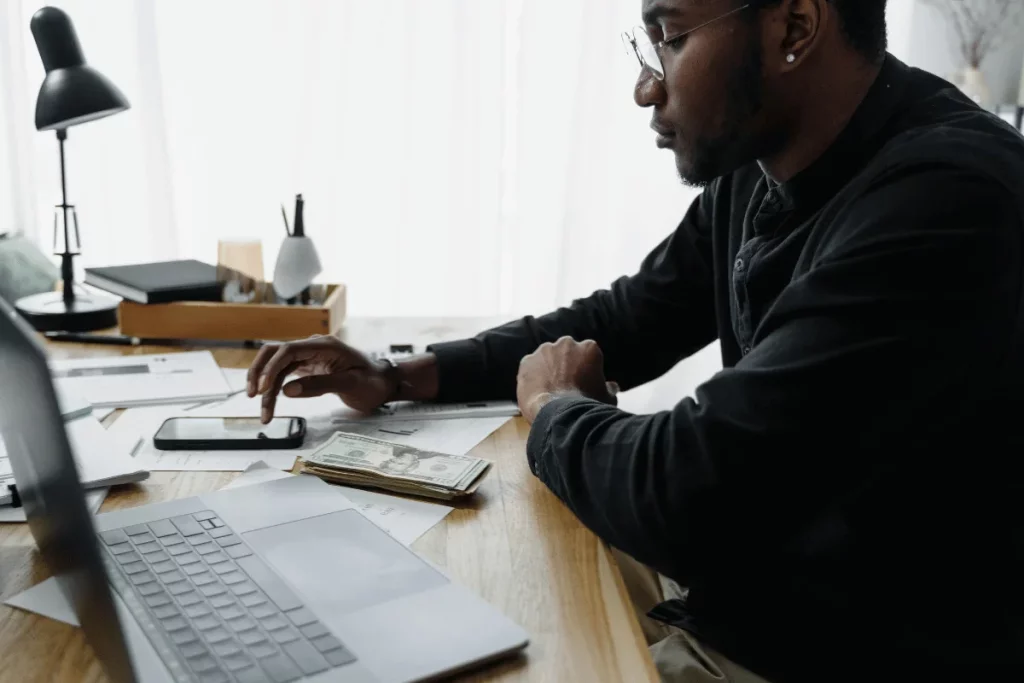 A man using a smartphone while seated at a desk with documents and cash stacked beside him.
