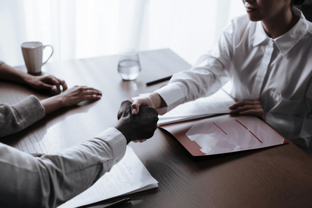 Two professionals in a handshake over a meeting table with documents.