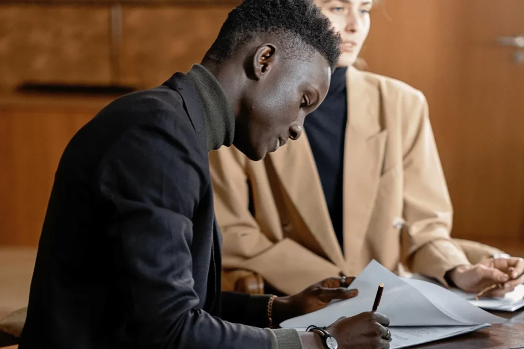 Two individuals engaged in a discussion at a table, one writing notes and the other gesturing, with documents spread out in front.