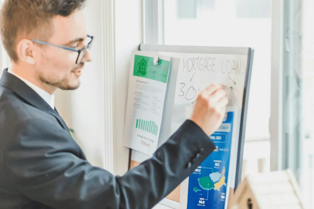 A man in a suit writes on a whiteboard, detailing mortgage loans and financial information, in a bright office setting.