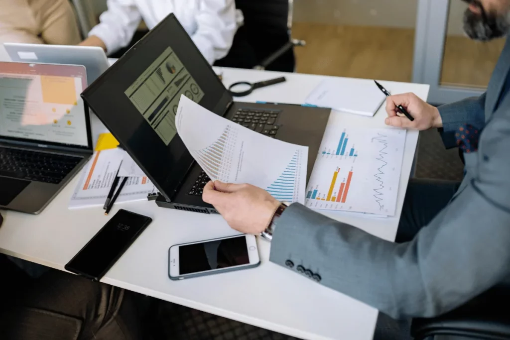A businessman reviewing reports and data on a laptop during a meeting, surrounded by documents and smartphones on a conference table.