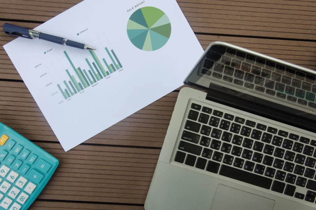 Sales report paper showing budget data and graphs, placed beside an open laptop on a wooden surface.