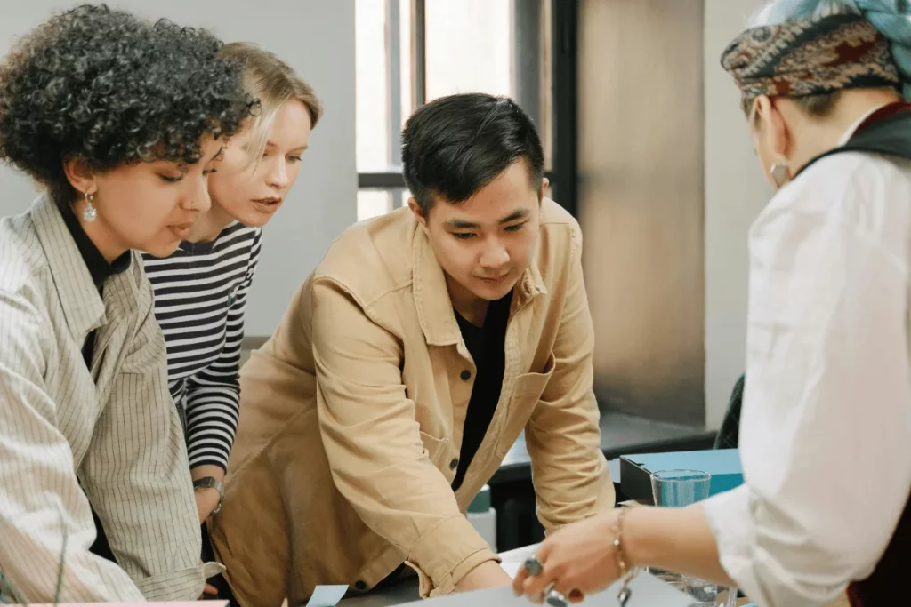 Four colleagues in a collaborative meeting, leaning over a table, engaging in teamwork and discussing project details in a bright workspace.