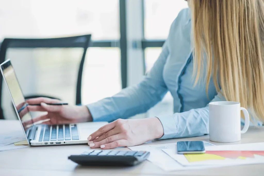 Person managing finances using a calculator and laptop on a white desk.
