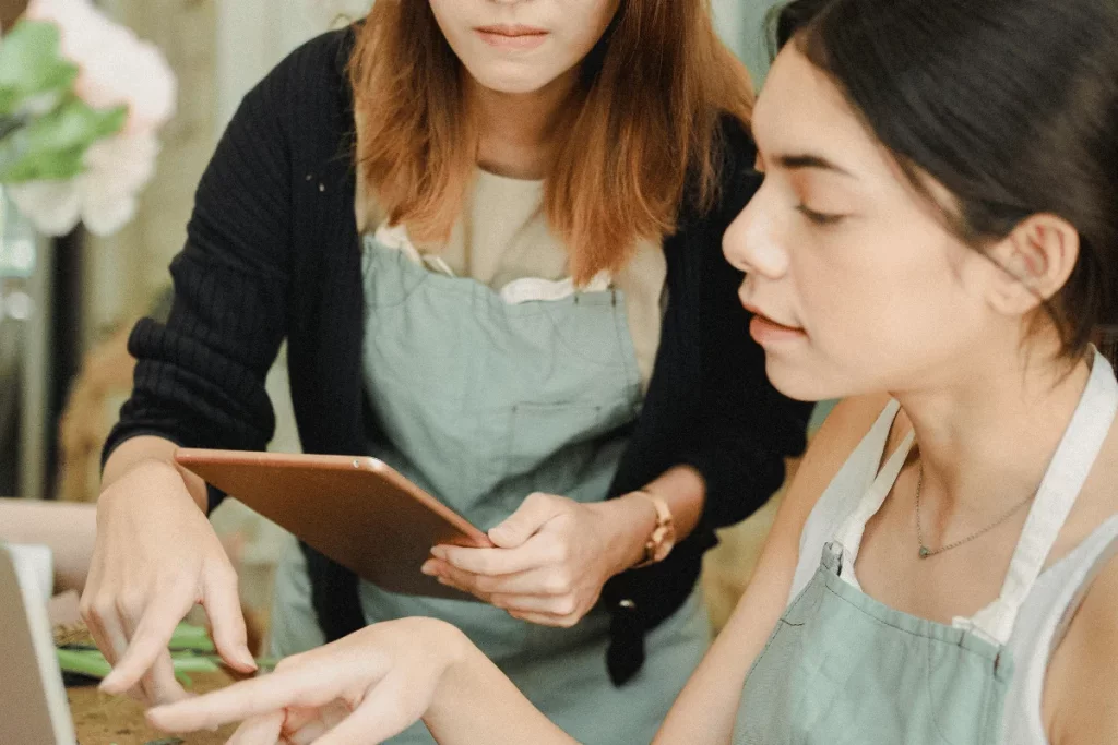 Two women in aprons working together, one holding a tablet and the other pointing.