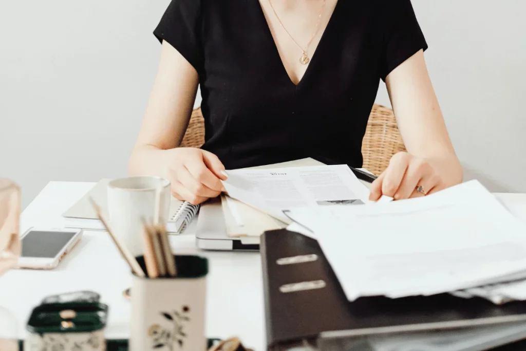 Person in a black shirt reviewing documents at a desk, with files, a notebook, and a smartphone nearby.