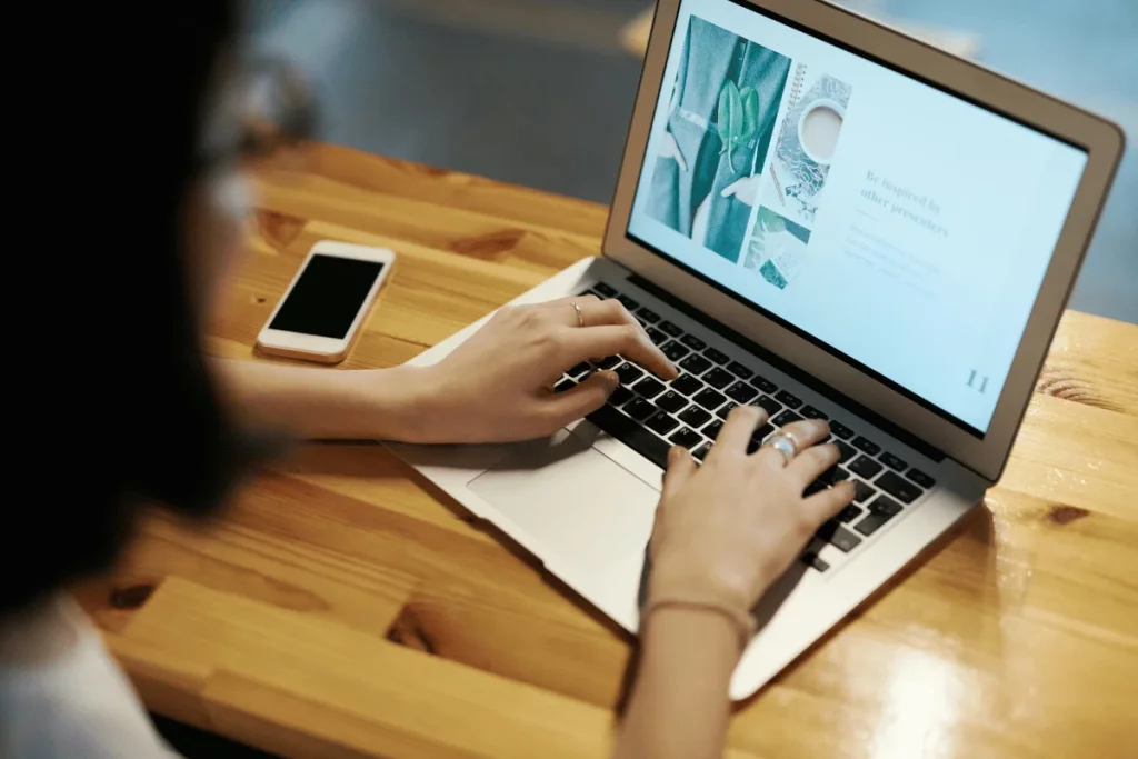 A person typing on a laptop with a design presentation visible on the screen, next to a white smartphone on a wooden table.