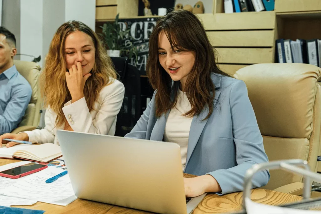 Two women are sitting at a table, smiling and looking at a laptop in a modern office setting.