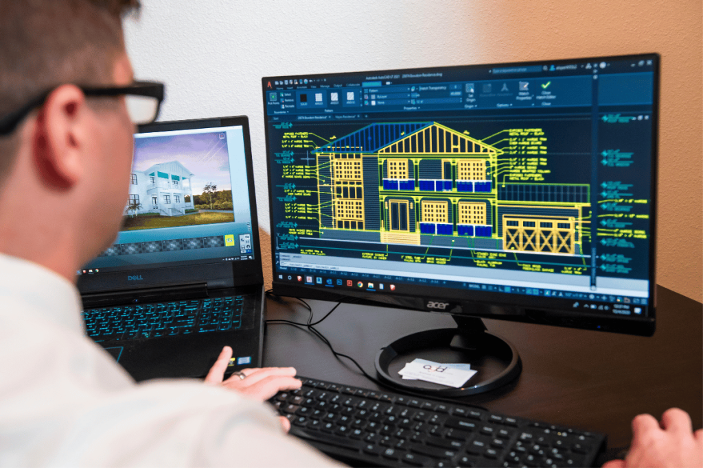 Architect reviewing a 3D house model and blueprints on dual computer screens.